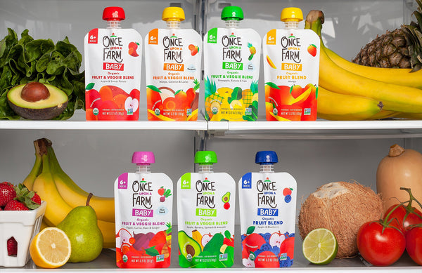 Various Once Upon a Farm baby food pouches on fridge shelves, surrounded by fresh fruits and veggies