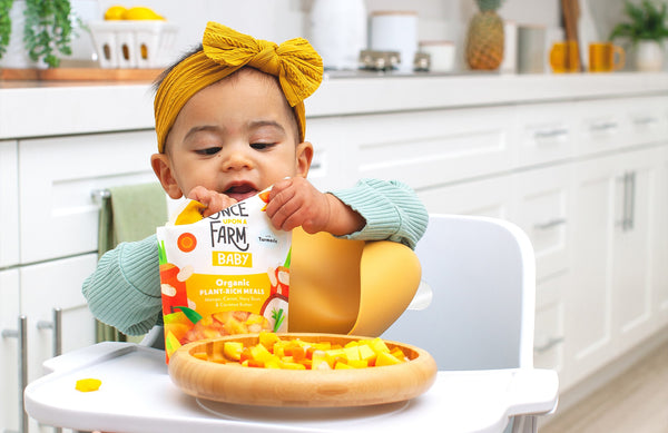 baby in highchair, holding a package of Once Upon a Farm frozen baby food, with the cooked meal on her plate