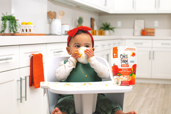 baby in highchair eating puréed baby food. Once Upon a Farm plant-rich meal packaging sitting on highchair tray