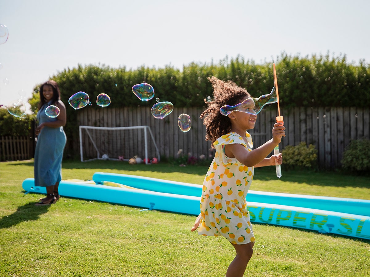 child blowing bubbles outside while her mom watches from a distance
