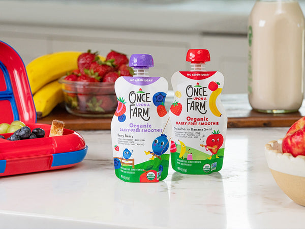 once upon a farm dairy-free smothies, on a counter, next to a lunchbox and some fresh fruit