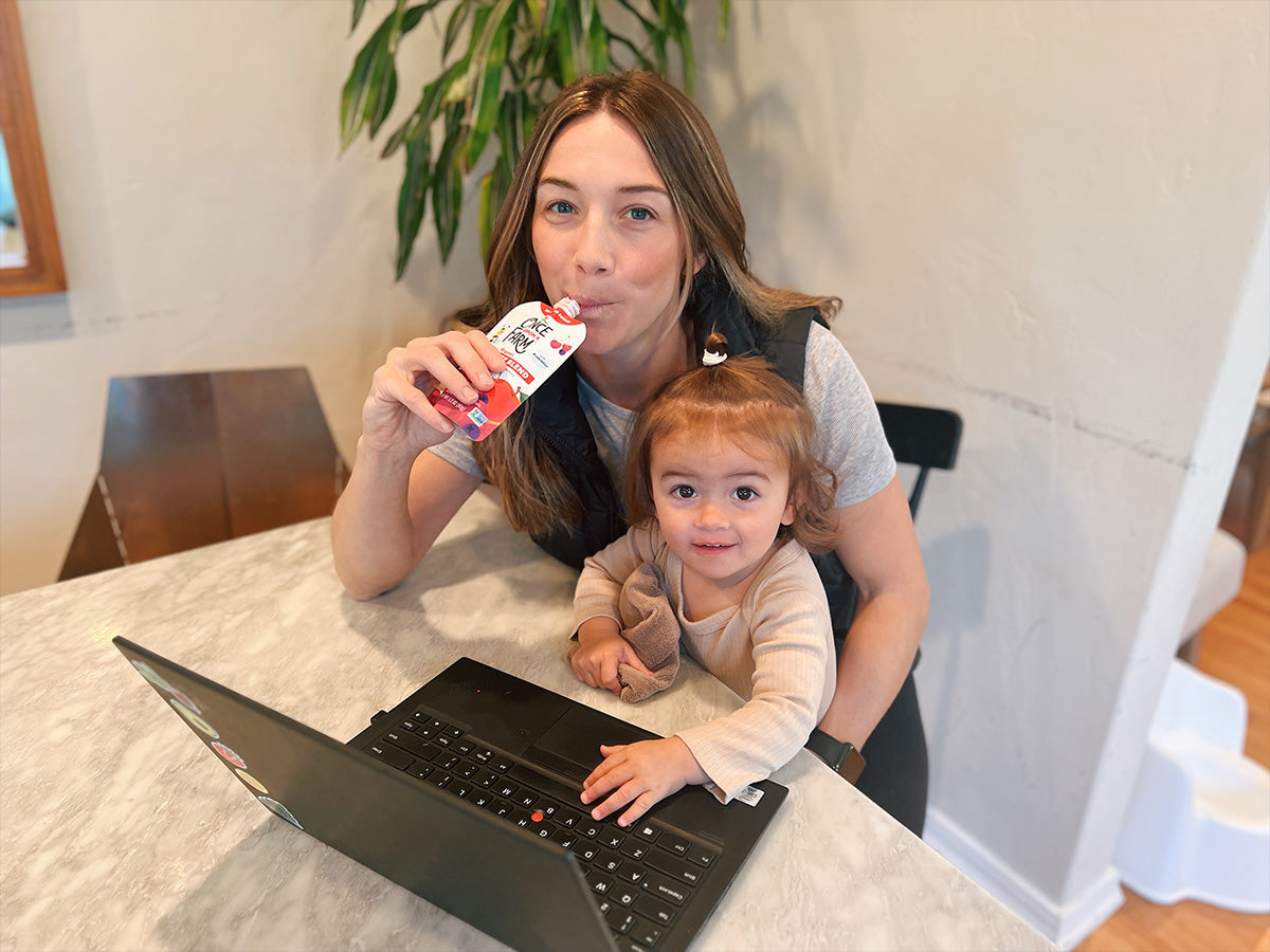 Once Upon a Farm Senior Director of Sales Kristin O'Connell, eating a pouch at her computer, while her daughter Maeve sits on her lap