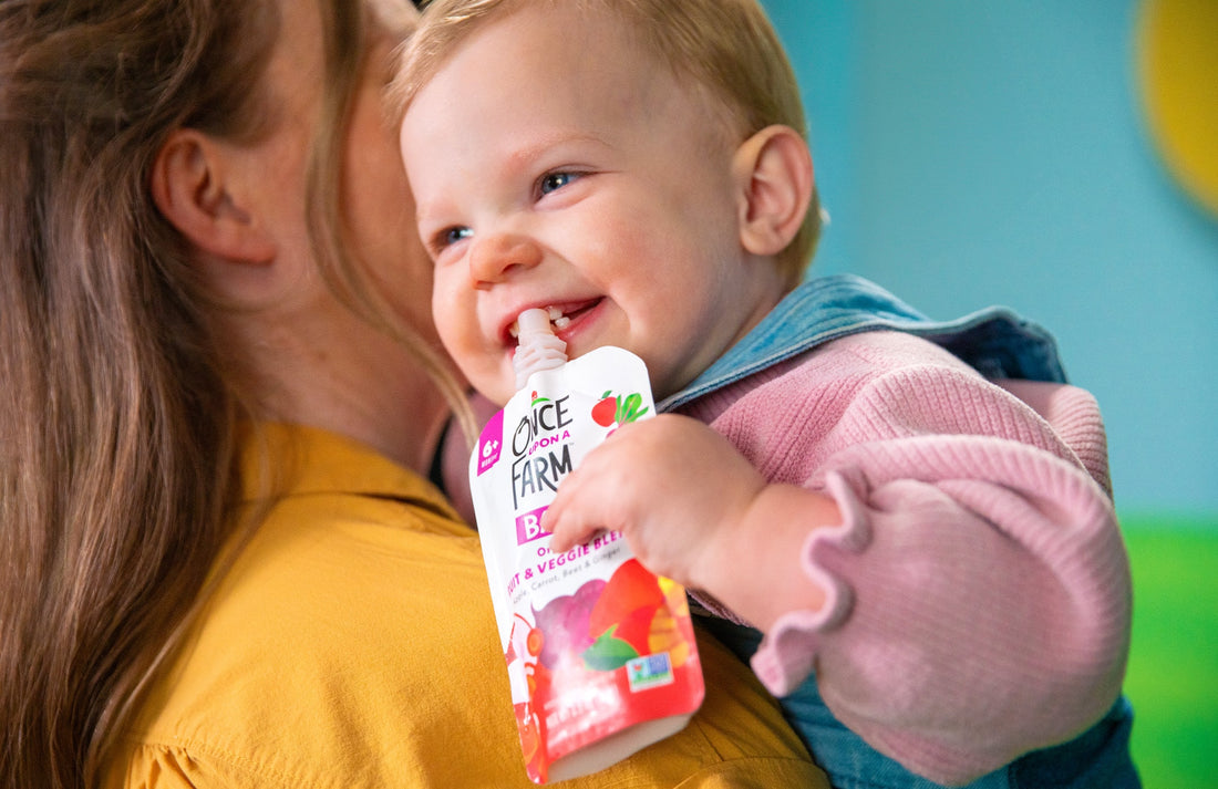 close up of a baby being held by mom. baby is holding a Once Upon a Farm pouch and smiling