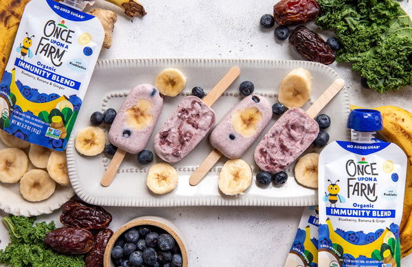 Frozen yogurt pops on a tray, surrounded by fresh fruit and veggies and Once Upon a Farm Blueberry, Banana & Ginger Immunity Blend pouches