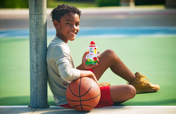 boy sitting on basketball court, leaning on a basketball, with a Once Upon a Farm smoothie pouch in his hand