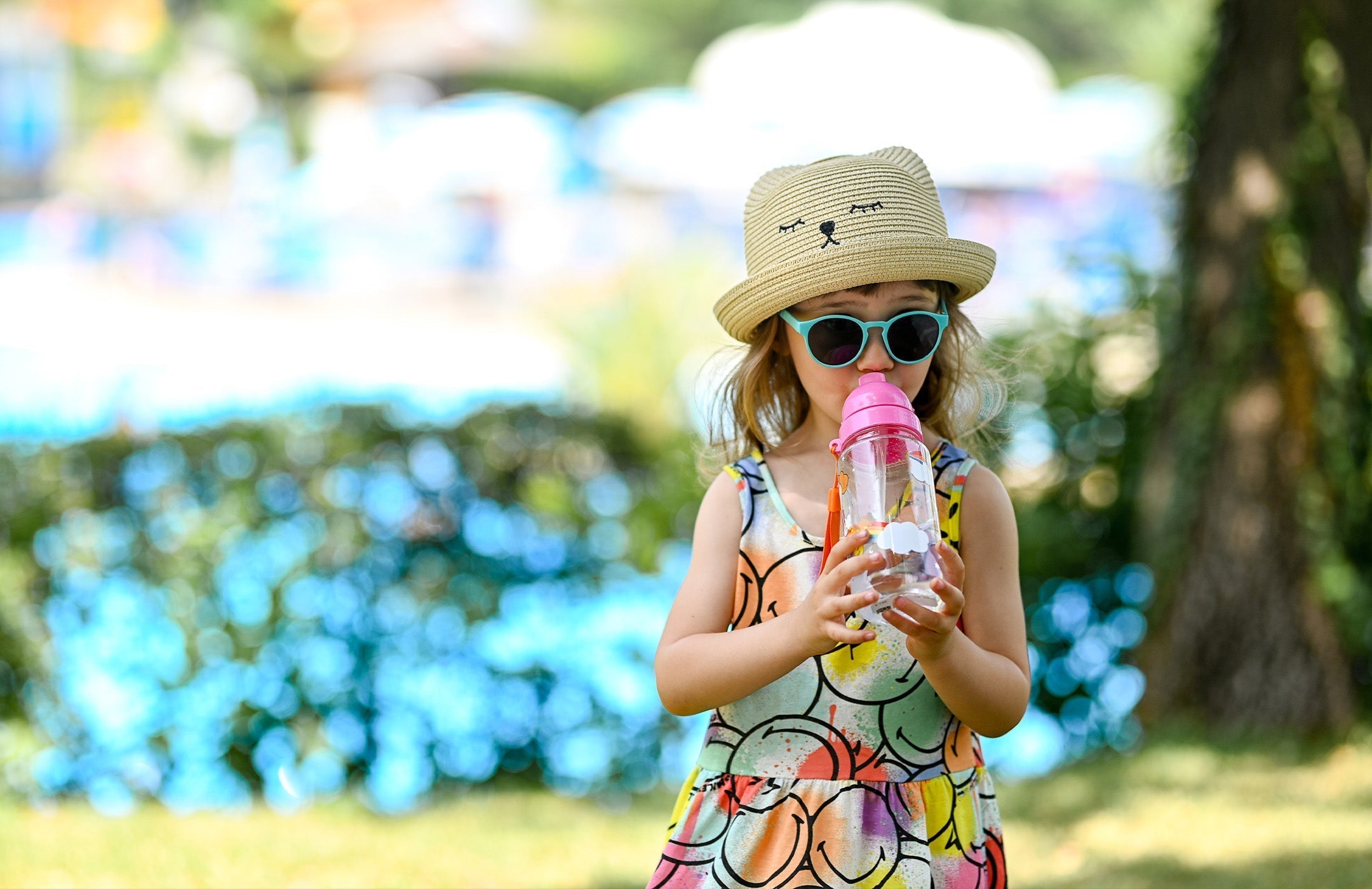 girl wearing a hat, sunglasses, and a sundress drinking from a water bottle outside on a sunny day