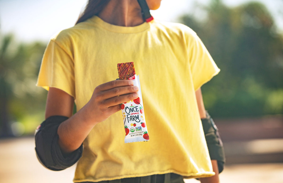 girl holding a Once Upon a Farm Refrigerated Oat Bar at a skate park. You can't see her face, but she is wearing a helmet and elbow pads