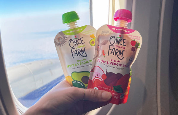 hand holding two Once Upon a Farm pouches in front of an airplane window in the sky