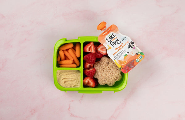 a bento-box style lunchbox with carrots, hummus, strawberries, a sandwich, and a Once Upon a Farm pouch