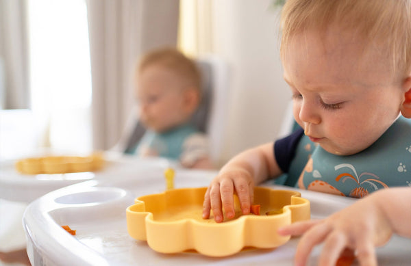 two babies in highchairs, eating food from plates