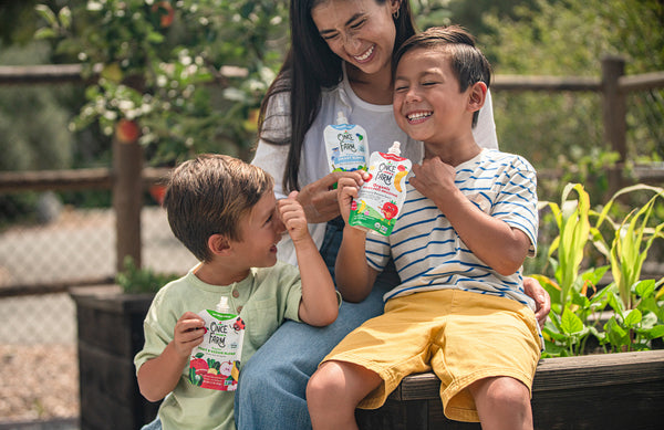 mom and two sons, sitting on a bench outside and smiling while each holding Once Upon a Farm snack pouches