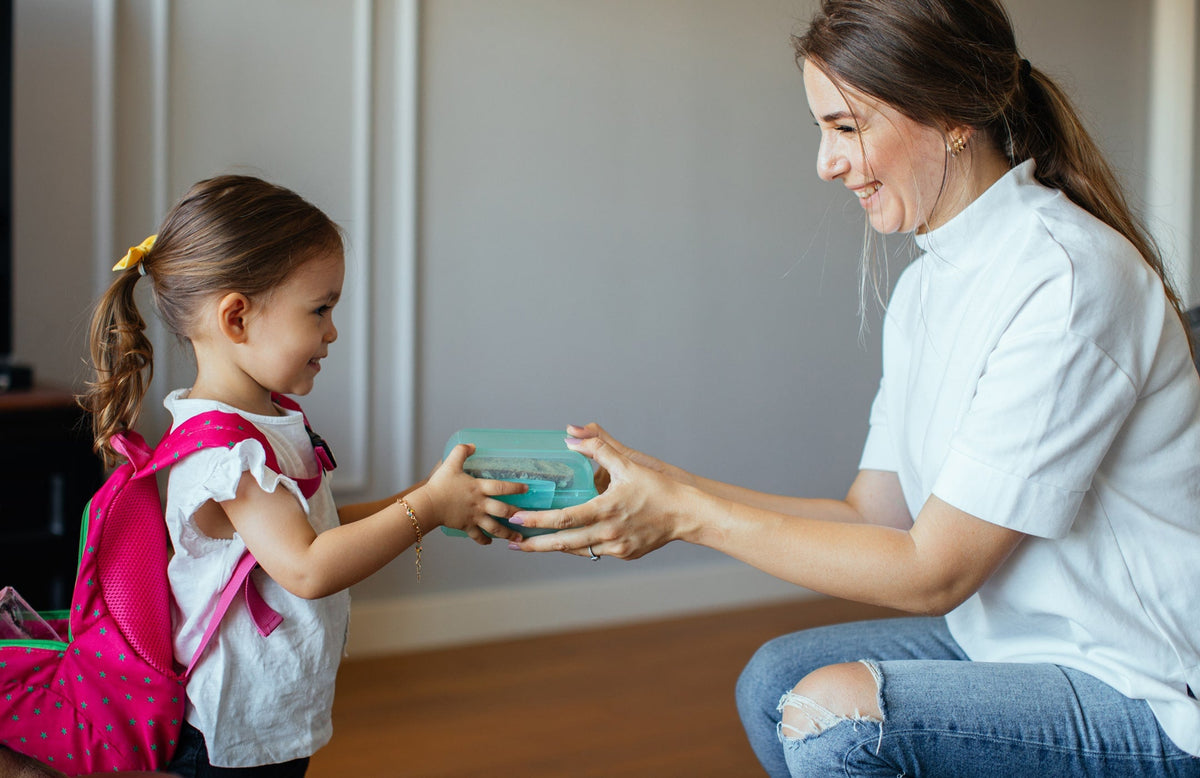 mom handing a lunchbox to a preschool-age girl with a backpack on