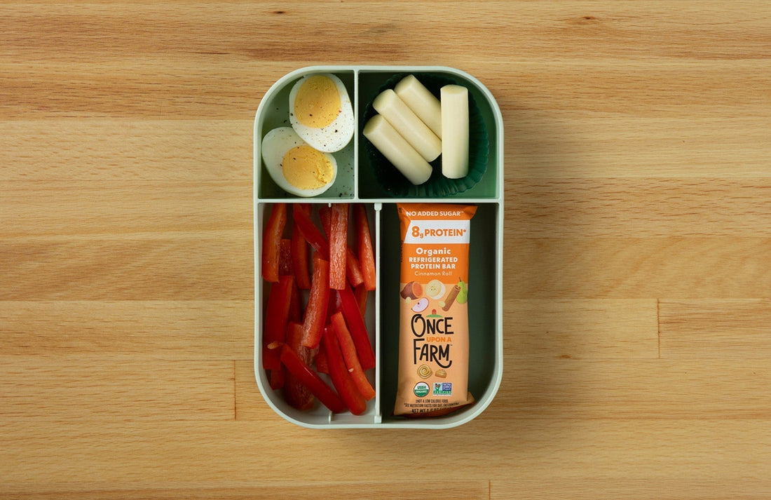 a bento-box style lunchbox with a hard-boiled egg, string cheese, red bell peppers, and a Once Upon a Farm Refrigerated Protein Bar
