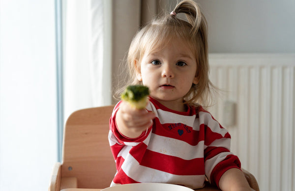 toddler girl in a high chair, holding out a broccoli floret toward the camera