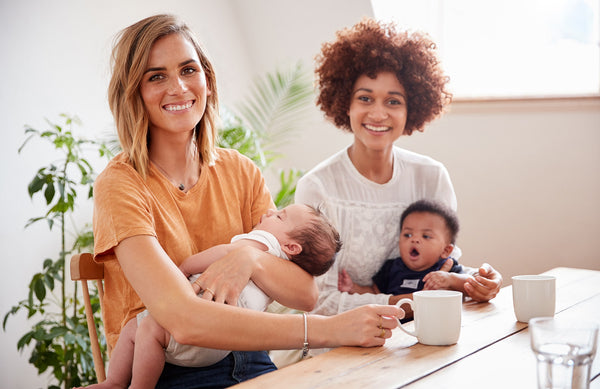 two moms, each holding a baby, having coffee together
